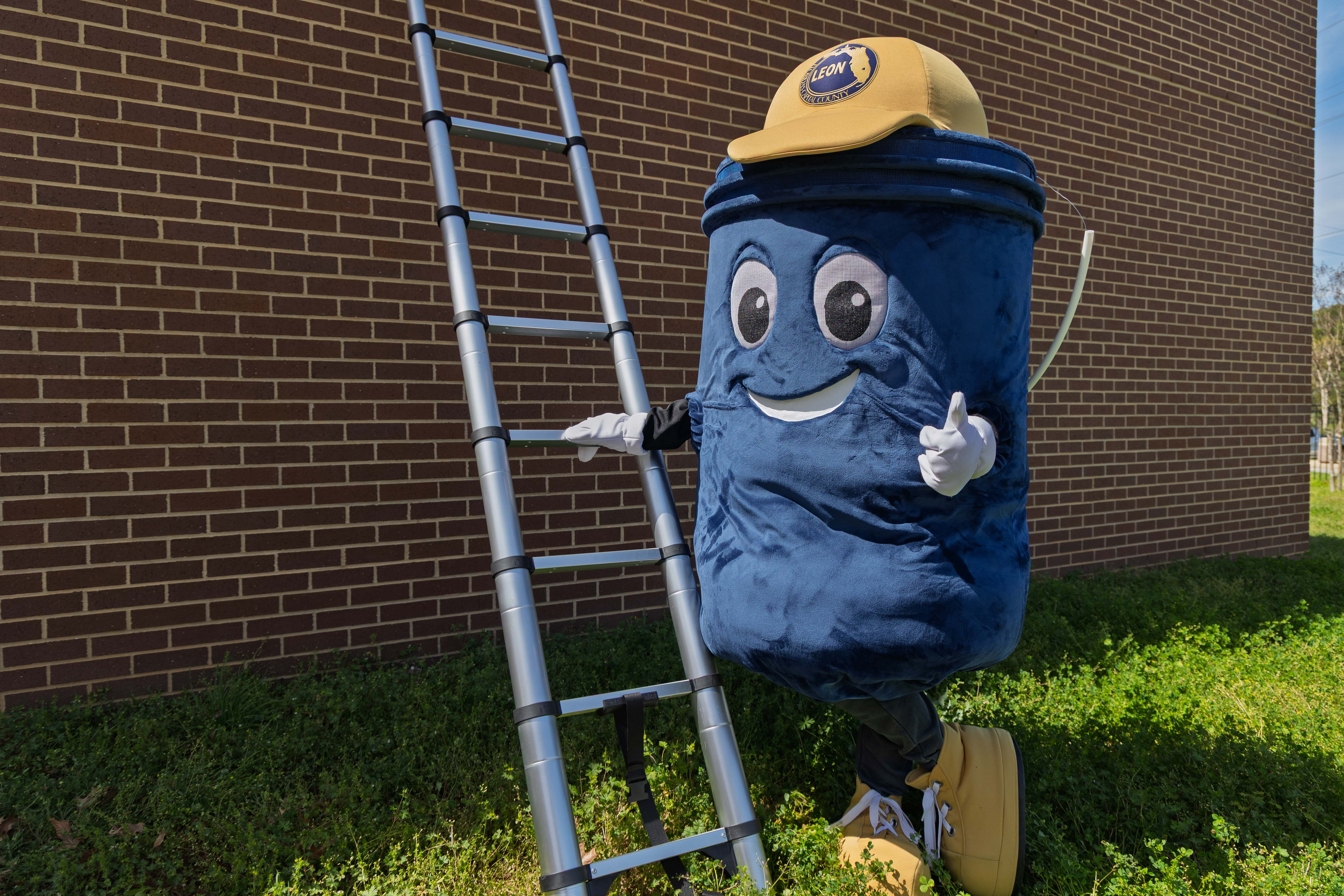 Smiling bucket mascot in a yellow cap gives a thumbs-up, standing next to a ladder along a brick wall,.